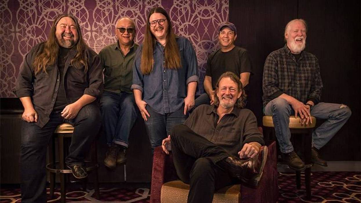 Six smiling men in casual clothes, some seated, some standing, in a group photo against a patterned wall.