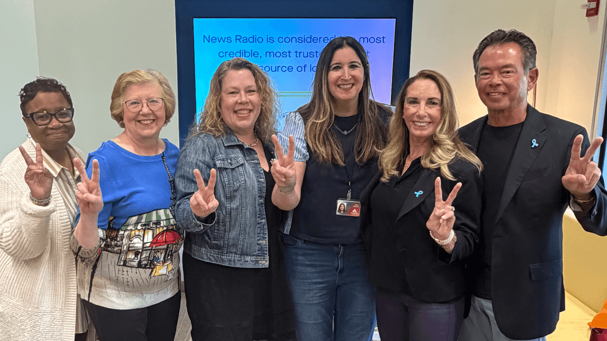 Six smiling people, four women and two men, making peace signs. Two wear blue ribbons.