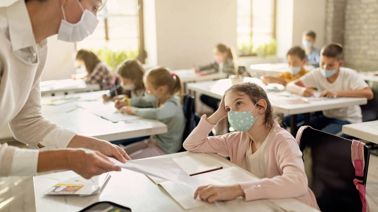 Small girl and her teacher wearing protective face masks while talking about exam papers during a class at elementary school.