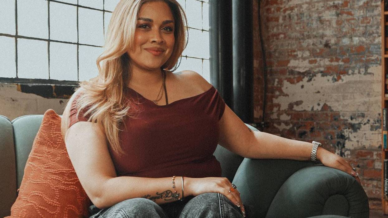 Smiling blonde woman on a green couch by a large industrial window and brick wall.