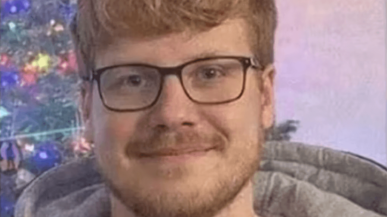 Smiling man with reddish hair, glasses, and beard in a grey hoodie, blurry Christmas tree background.