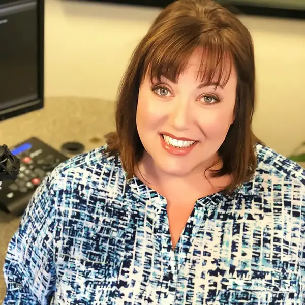Smiling woman with brown hair in a blue patterned shirt at a radio studio.