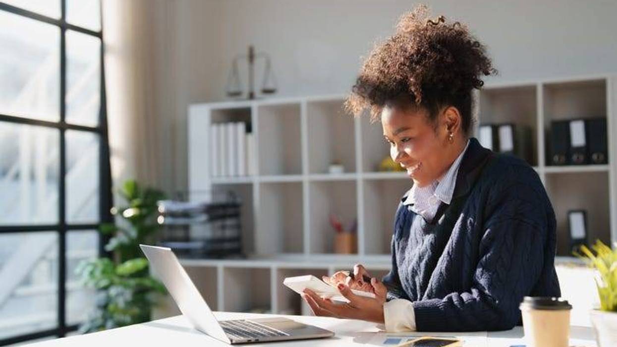 Smiling young adult woman concentrating on financial calculations using a calculator and laptop, working on business accounting and taxes in a professional environment