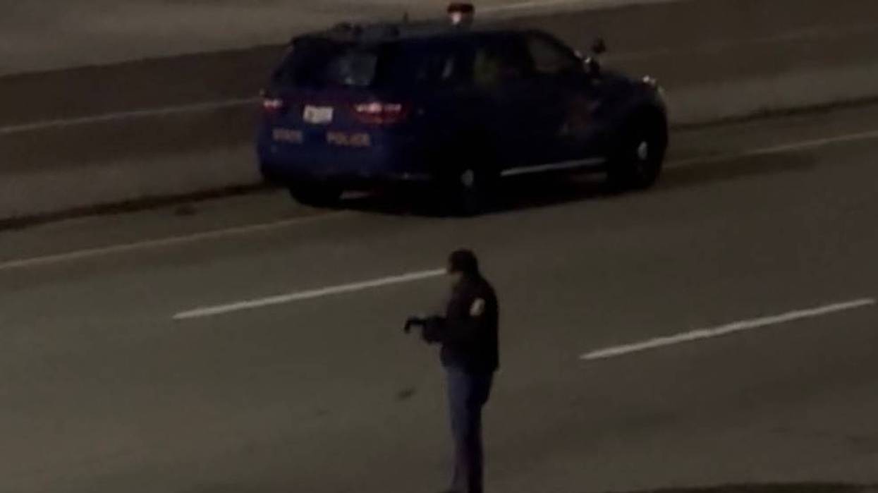 State police officer holding a rifle stands on a dark highway with their cruiser behind them.