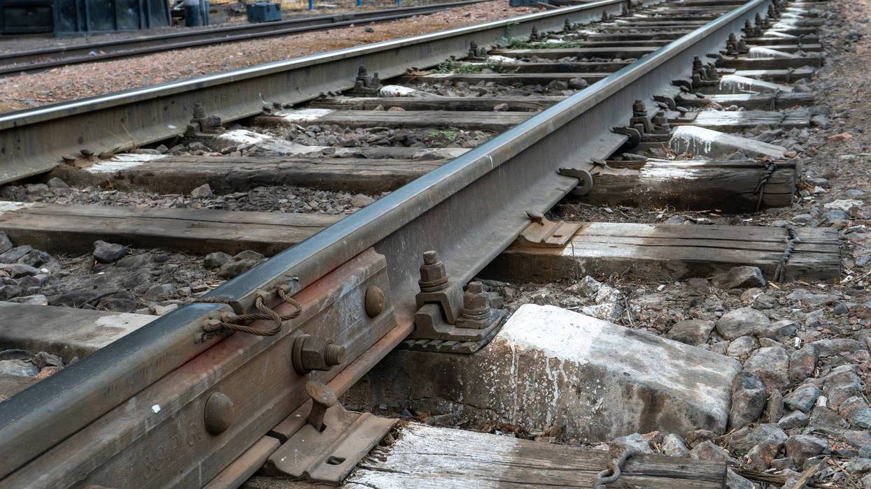 Steel railway tracks mounted on weathered wooden sleepers with metal fasteners and bolts supported by coarse gravel ballast, forming durable foundation for train movement in industrial environment