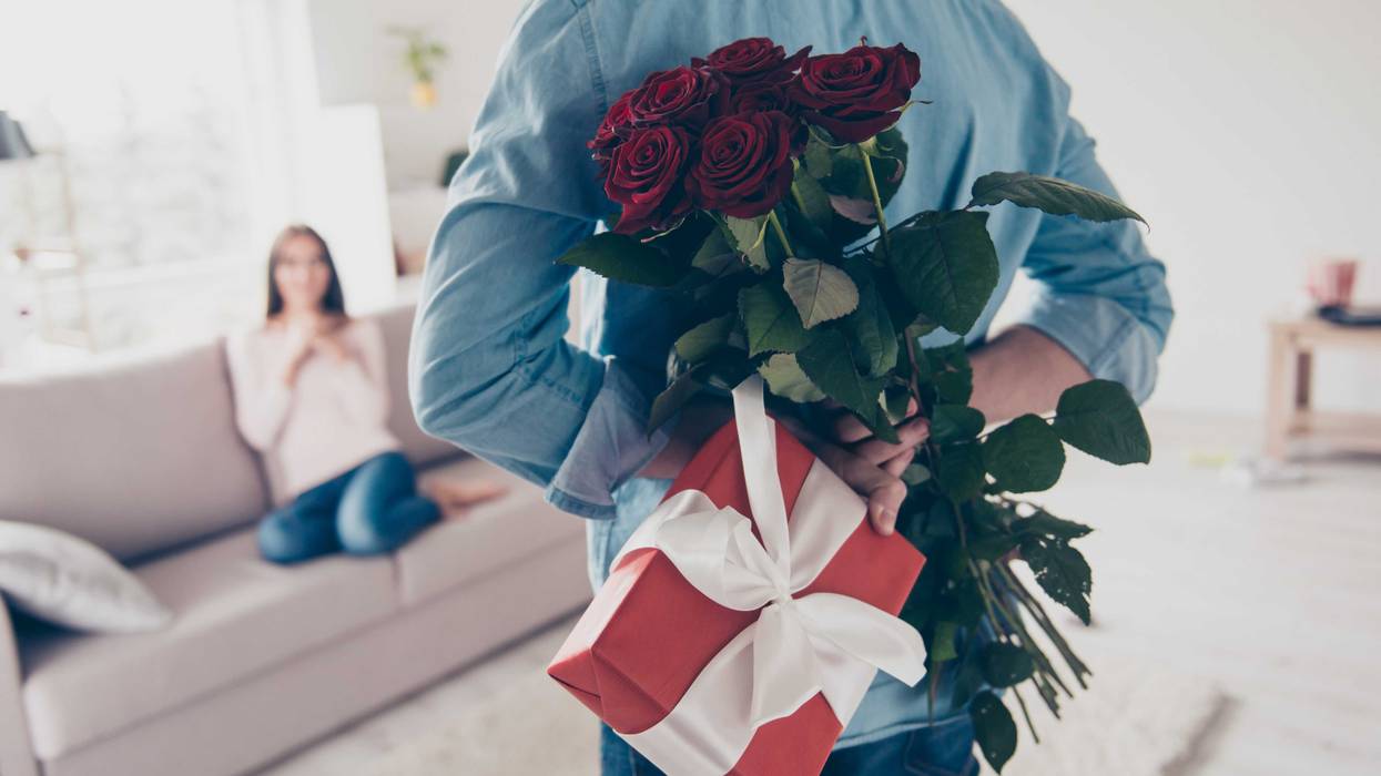 stock image. woman sits on couch, man stands in front of camera facing her holding roses and a gift behind his back for valentine's day