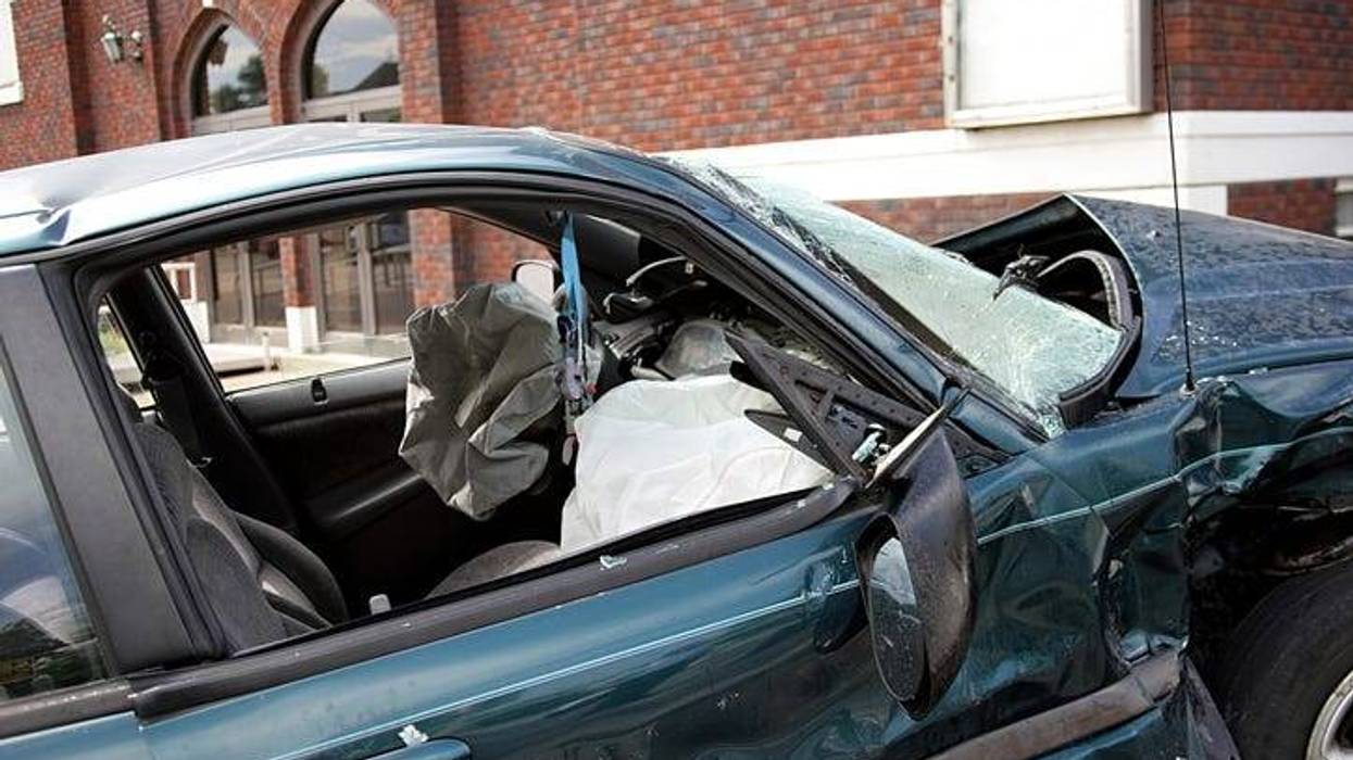 Stock photo of a car crashed near a brick building. The windows are broken, hood is smashed and airbags have deployed.