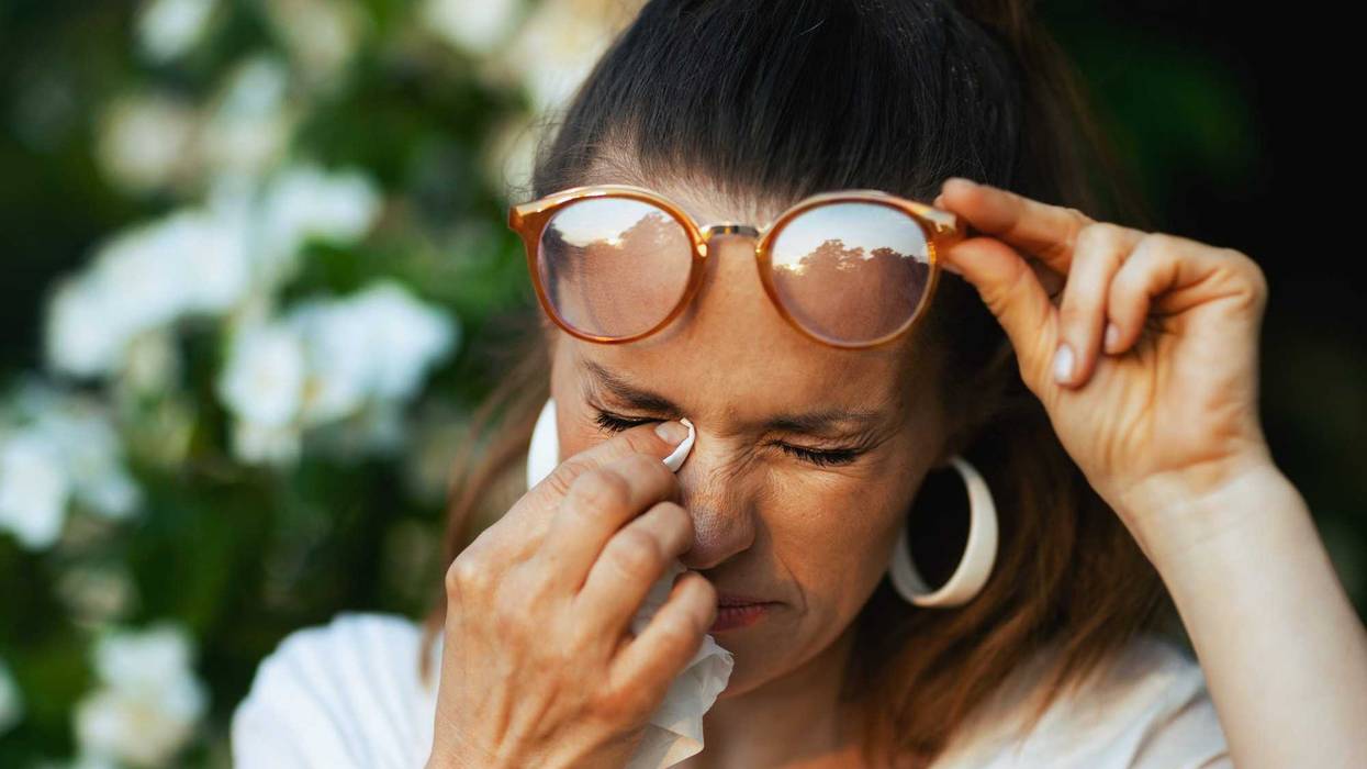 Summer time. sad modern 40 years old woman in white shirt with handkerchief and eyeglasses has an allergy attack near flowering tree.