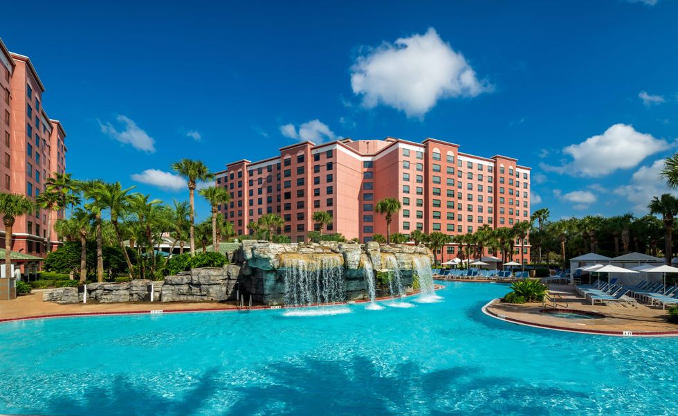 Sunny tropical resort with large peach-colored hotel, pool, rock waterfalls, and palm trees.