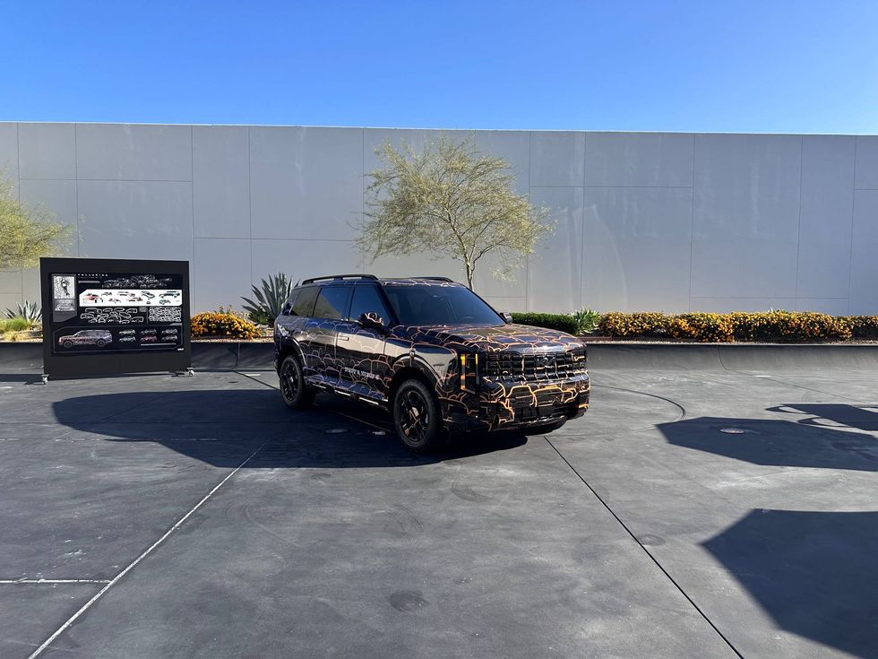 SUV with a striking black and orange camouflage pattern parked on a sunny concrete lot, near a display board and surrounded by plants and a modern wall.
