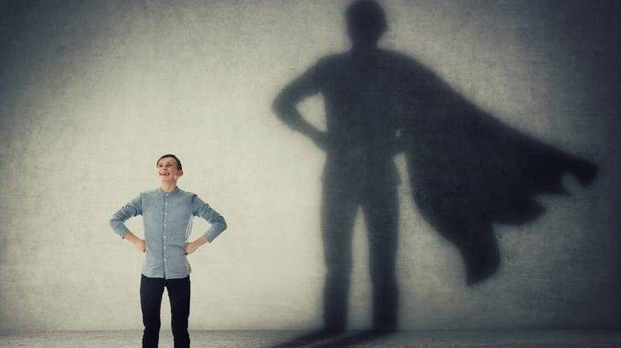 Teenager, with arms on hips smiling confidently, casting a superhero with cape shadow on the wall.