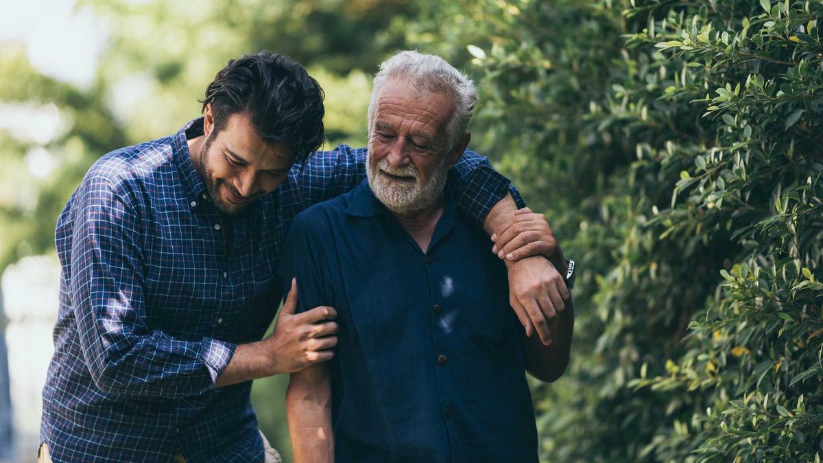 The old man and his son are walking in the park. A man hugs his elderly father. They are happy and smiling