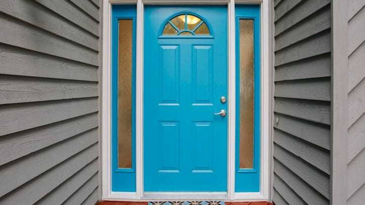 This image showcases a bright blue front door with two narrow side panels of frosted glass, a silver handle, and a welcome mat on a red wooden porch floor, surrounded by gray siding walls, creating a welcoming entrance to the home.