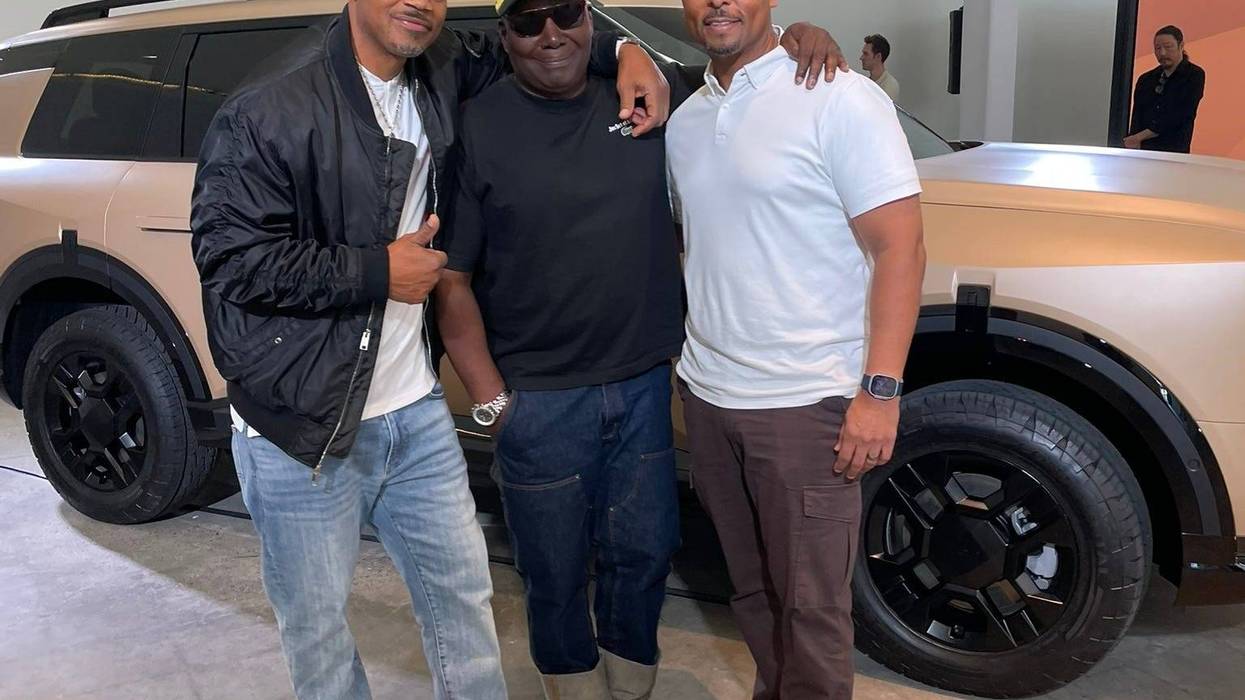 Three men stand smiling in front of a stylish SUV inside a modern showroom. One wears a black jacket and cap, another a yellow cap, and the third a white shirt.
