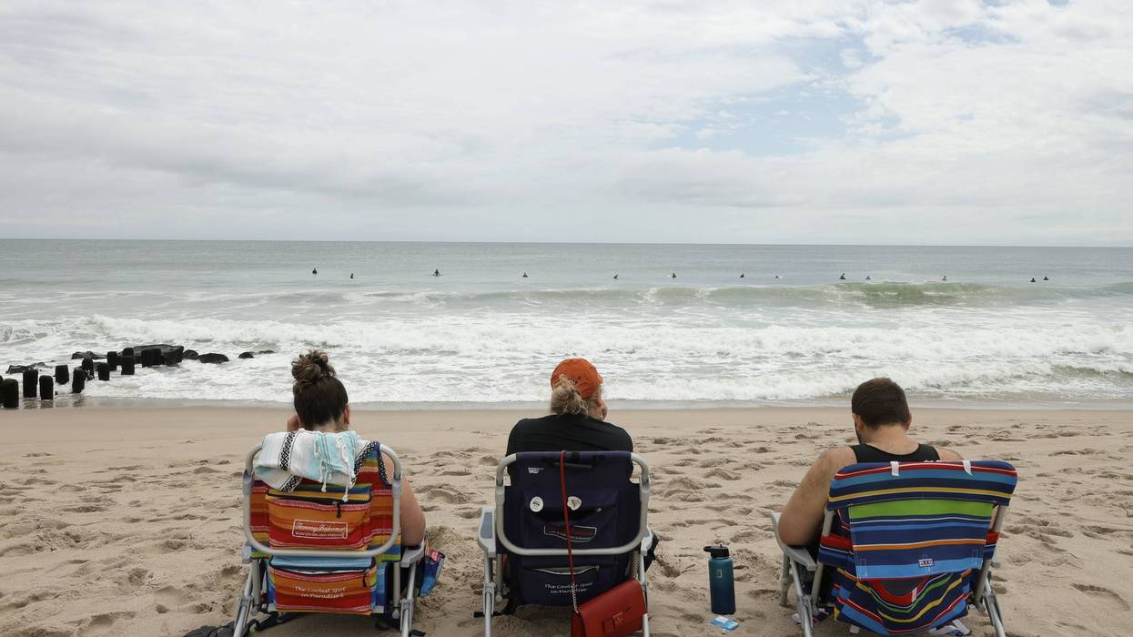 Three people sit in beach chairs on sand, watching surfers in the ocean under a cloudy sky.