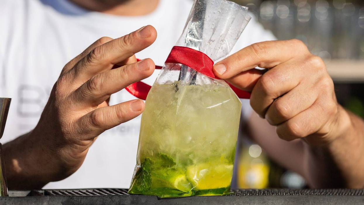 Two bartender's hands tie a red ribbon to a plastic take-out can containing a lime, lemon and mint mojito. Above a bar (pub) outside, it's daytime.