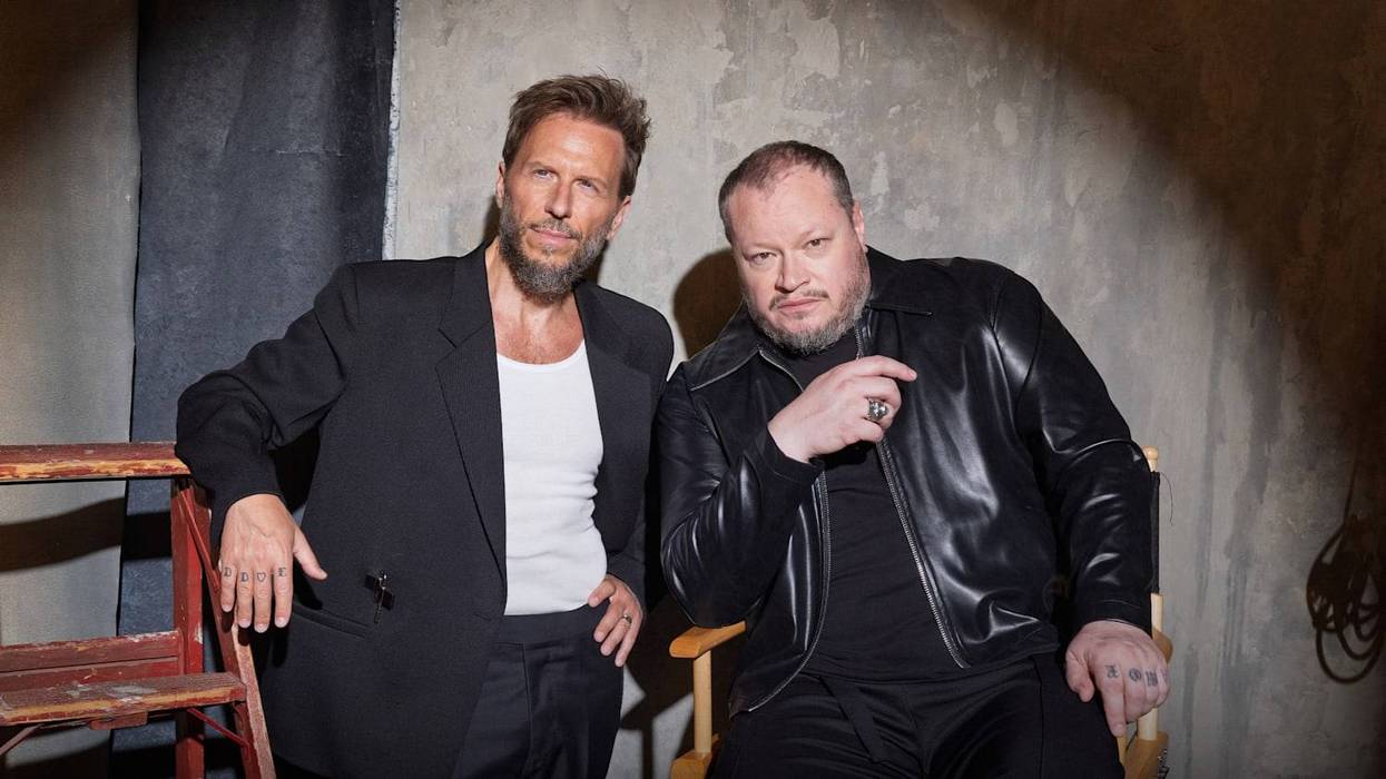Two bearded men, one in a black blazer, the other in a black leather jacket, against a textured wall.