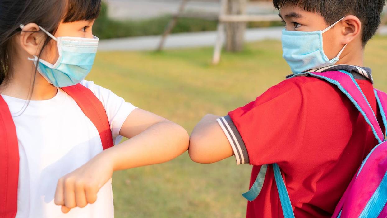 two children wearing backpacks and face masks elbow bump