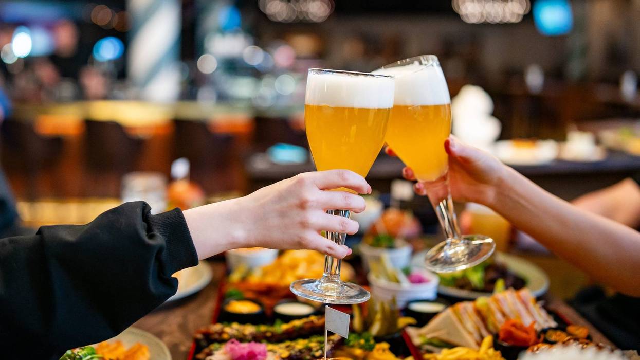 Two hands clinking beer glasses in a cozy indoor setting. Assorted food items are visible on a wooden table in the blurred background - stock photo