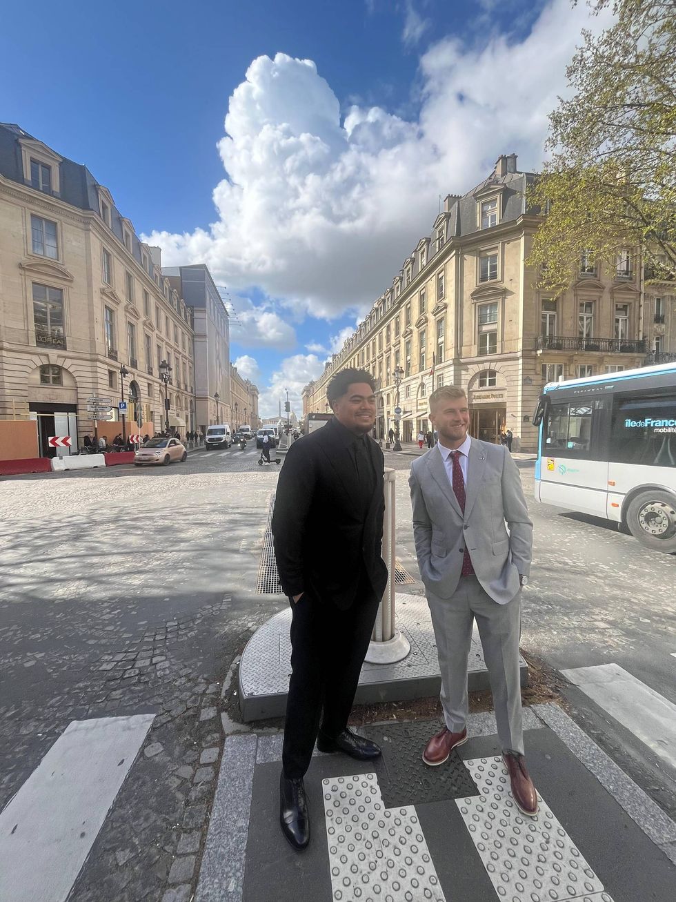 Two men in suits stand on a sunny Paris street corner with historic buildings and a bus.