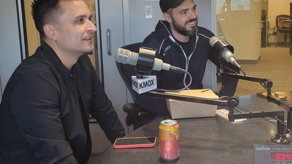 two men sit at kmox microphones they're both wearing black jackets and one wears a black hat that says balkan