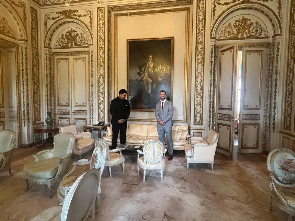 Two men stand in a grand, gilded room with a large portrait and ornate antique furniture.