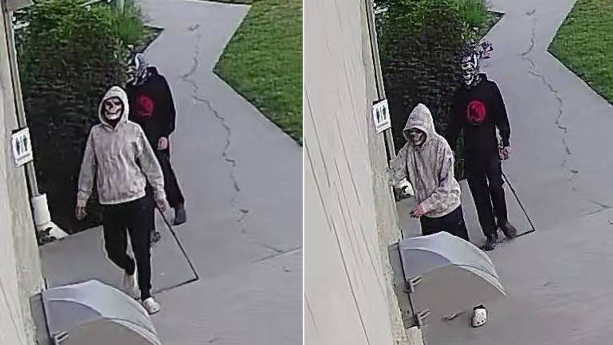 Two people in skull masks and hoodies walk with sticks on a path; an umbrella is on the ground.