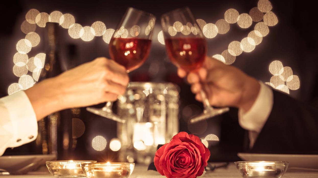 Two people toast with wine in the background of a table set with a rose and candles