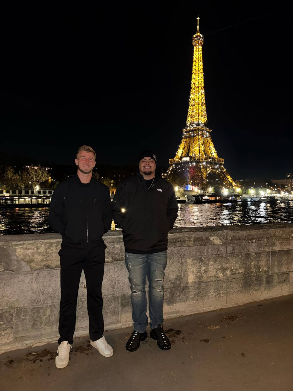 Two smiling men stand by the Seine River with the brightly lit Eiffel Tower at night.