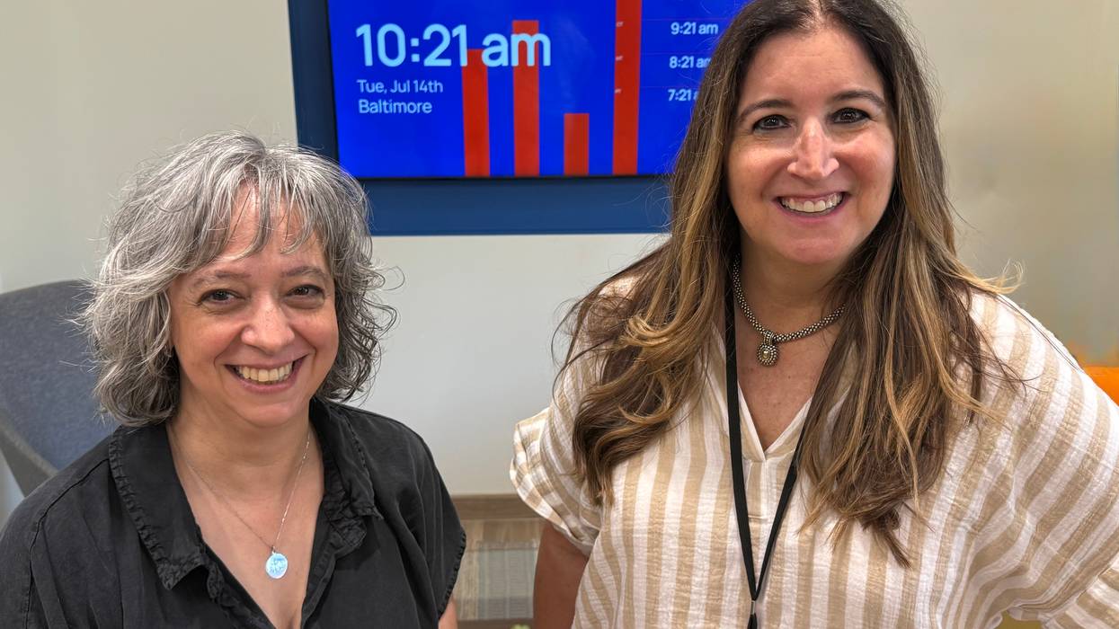Two smiling women, one grey-haired, one brown-haired, by a digital screen showing 10:21 AM.