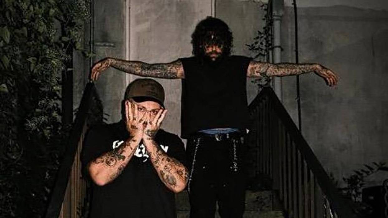 Two tattooed men on dark steps. Capped man covers face; messy-haired man has outstretched arms.