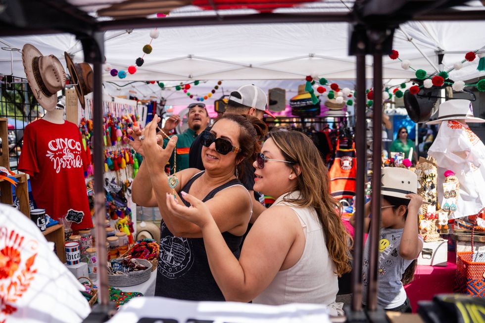Two women examine jewelry at a Mexican-themed outdoor market with colorful crafts and decorations.