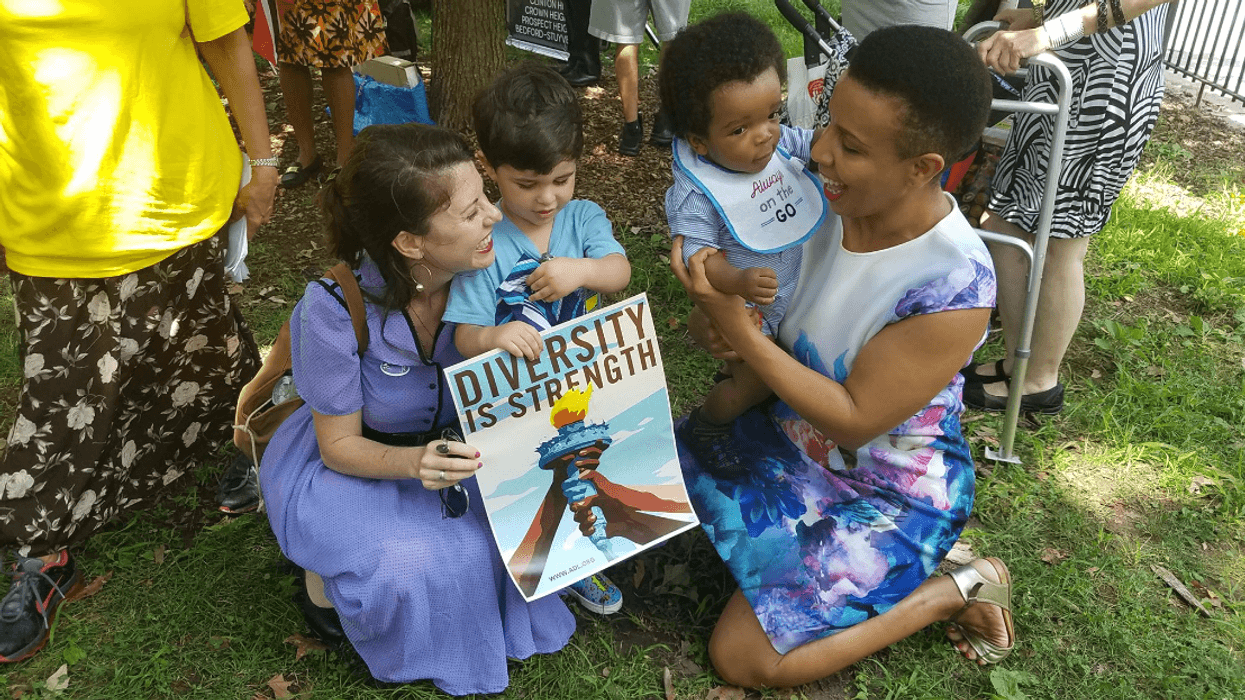 Two women, one Black and one Jewish, kneel together with young children and a sign that reads "diversity is our strength" at the One Crown Heights Neighborhood Festival.