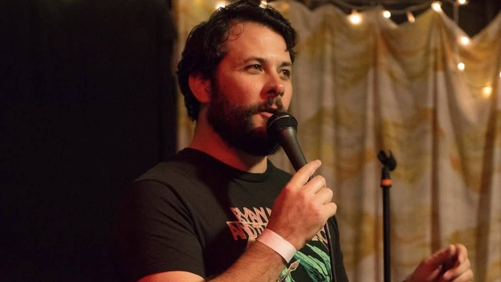 \u200bJoe Cardosi speaking into a microphone on stage, lit by string lights.