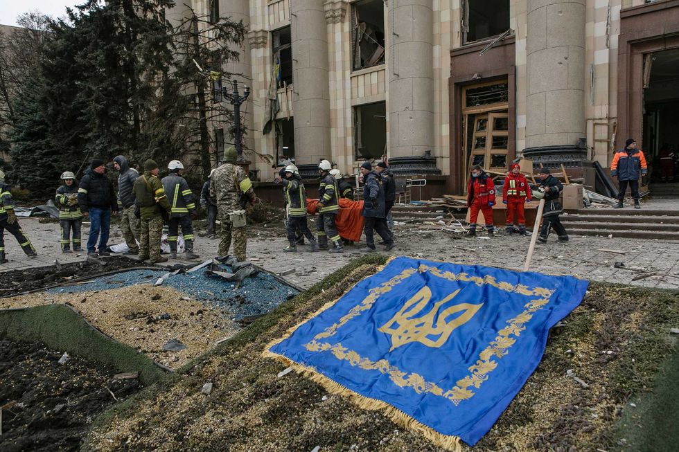 Ukrainian emergency service personnel walk past a Ukrainian coat of arms while carrying a body of a victim following shelling of the City Hall building in Kharkiv, Ukraine, Tuesday, March 1, 2022