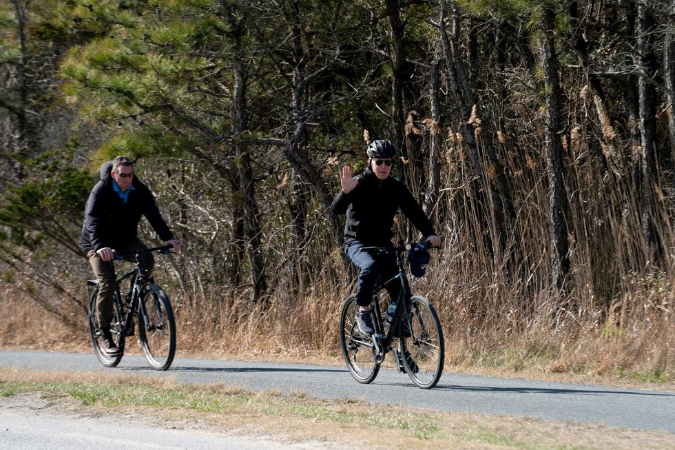 US President Joe Biden (R) waves while riding his bike through Cape Henlopen State Park in Rehoboth Beach, Delaware, on March 20, 2022.