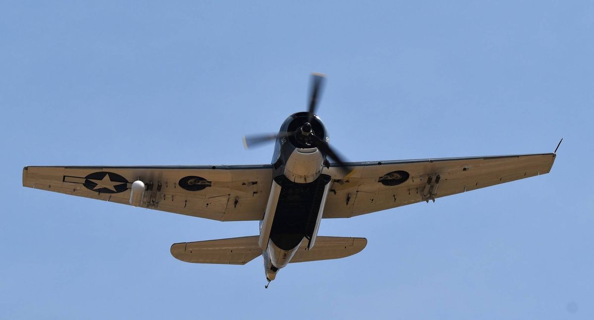 Vintage F4F Wildcat fighter plane flying overhead, underside visible against a blue sky.
