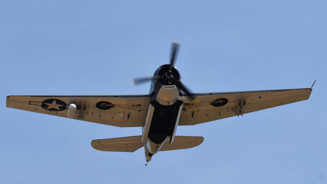 Vintage F4F Wildcat fighter plane flying overhead, underside visible against a blue sky.