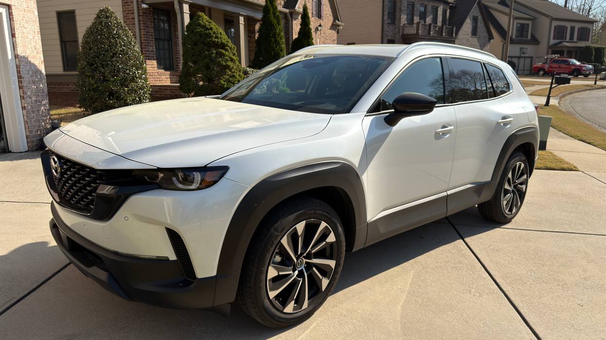 White Mazda CX-50 SUV parked on a sunny driveway with suburban houses in the background.