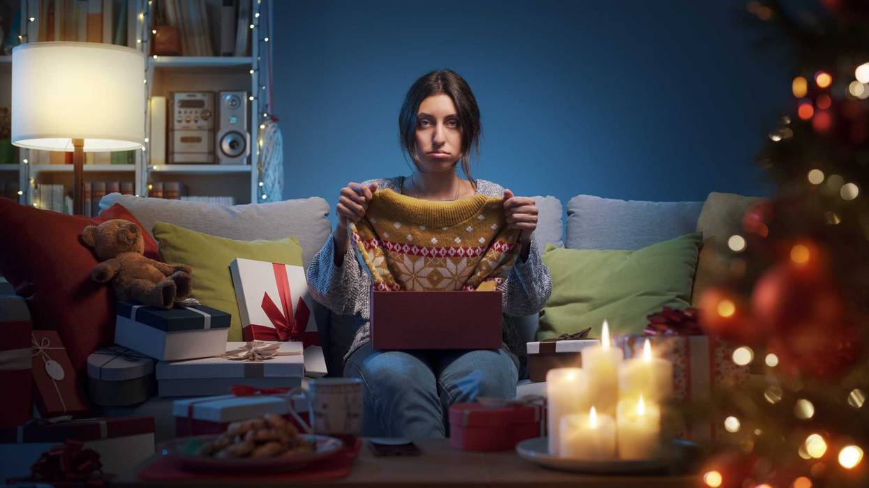 Woman holding sweater with unimpressed face next to a Christmas tree.