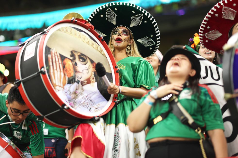 Woman in green Catrina makeup & sombrero sings next to a drum with Catrina art.