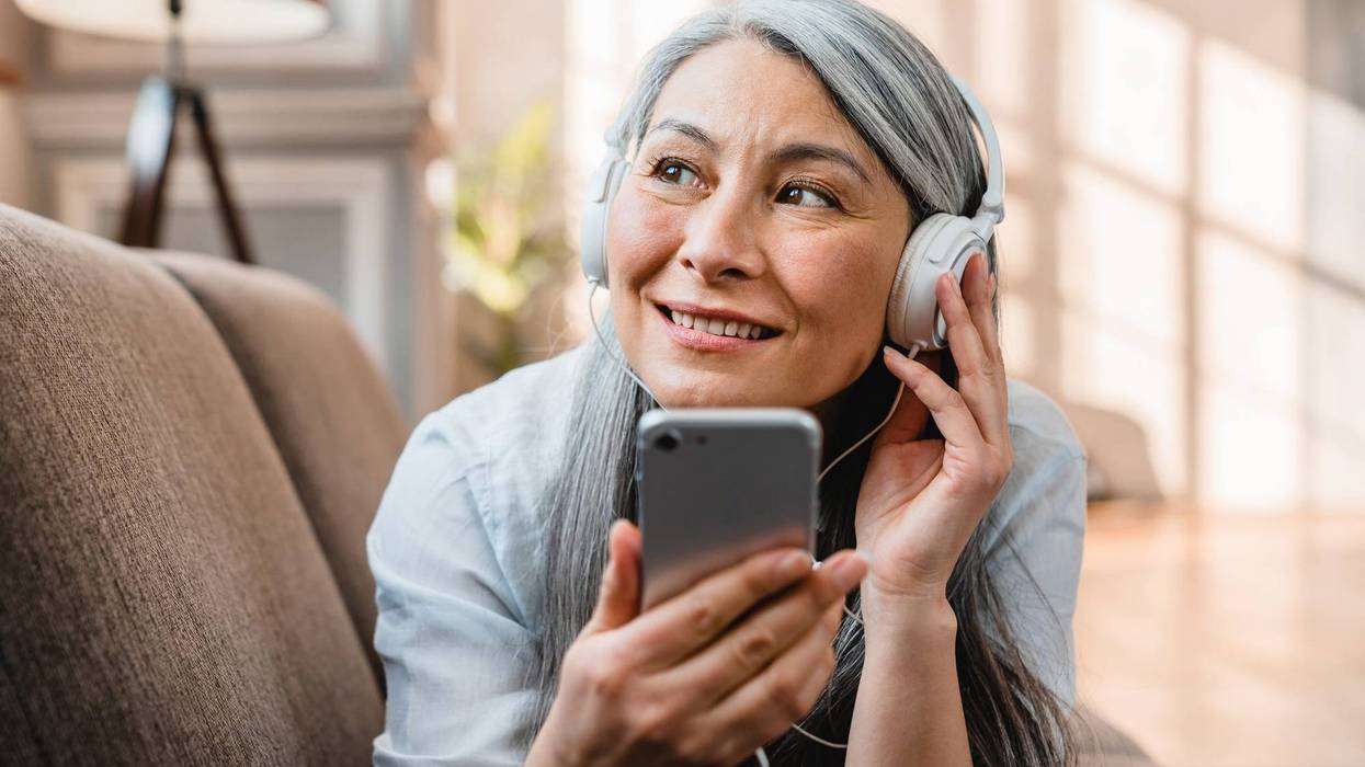 Woman lying belly down on couch holding a cellphone and enjoying audio on her headphones.