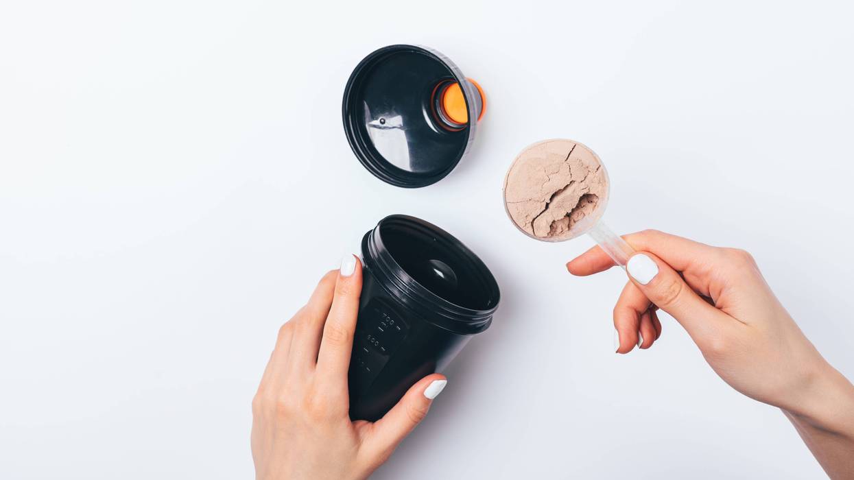 Woman's hands putting scoop of chocolate protein powder into black plastic shaker to prepare sports drink for muscle growth, top view on white table.