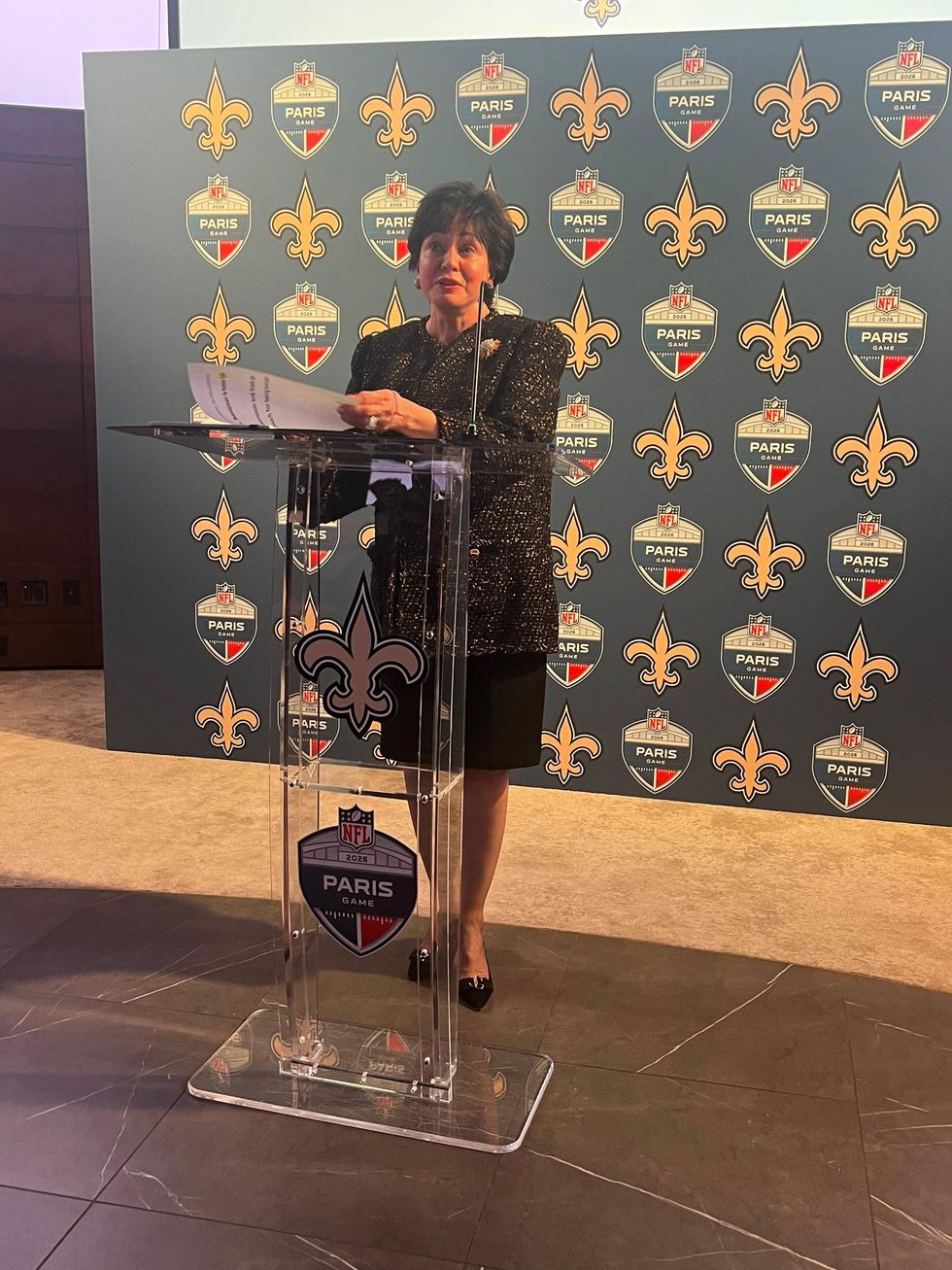 Woman speaking at a podium with NFL Paris Game and New Orleans Saints logo background.