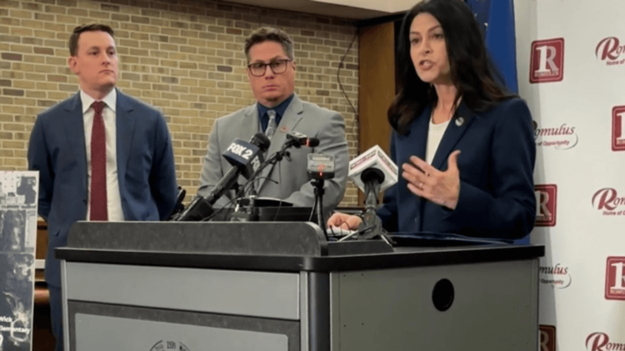Woman speaking at a press conference podium with two men, microphones, and a Romulus banner.