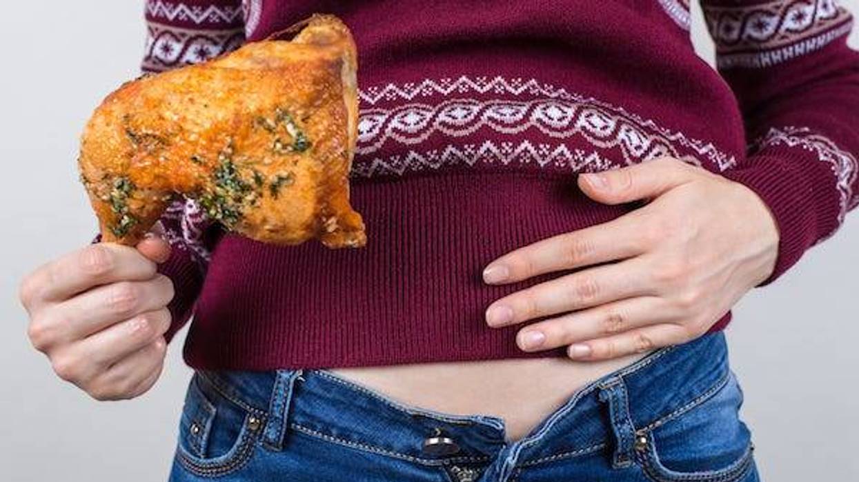 Woman wearing a holiday sweater with her jeans unbuttoned patting her belly while holding a rotisserie chicken