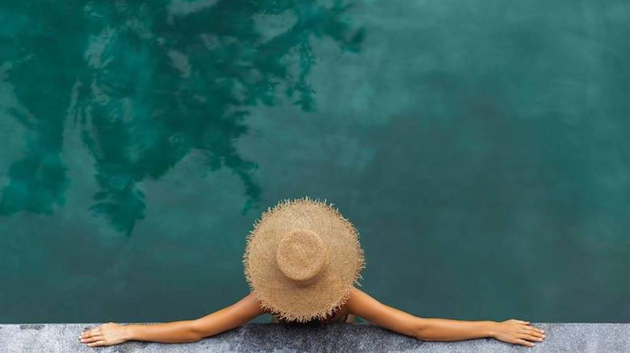 Woman wearing straw hat relaxing in swimming pool with arms stretched on wooden deck