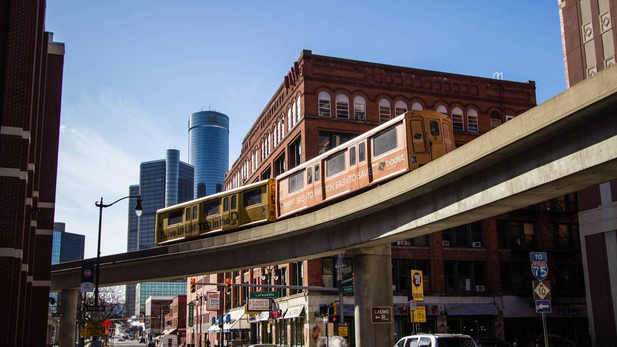 Yellow elevated train on tracks above a city street, with brick buildings, skyscrapers, and blue sky.