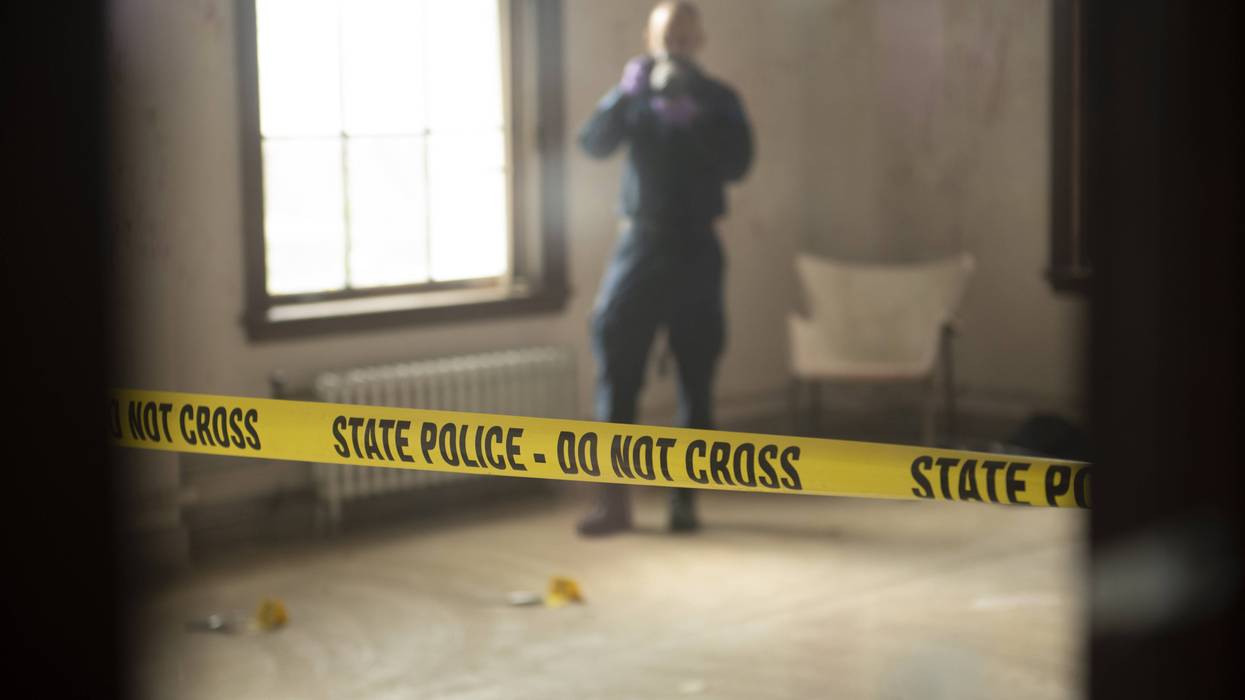 Yellow State Police tape in foreground; blurry investigator and evidence markers visible in background.