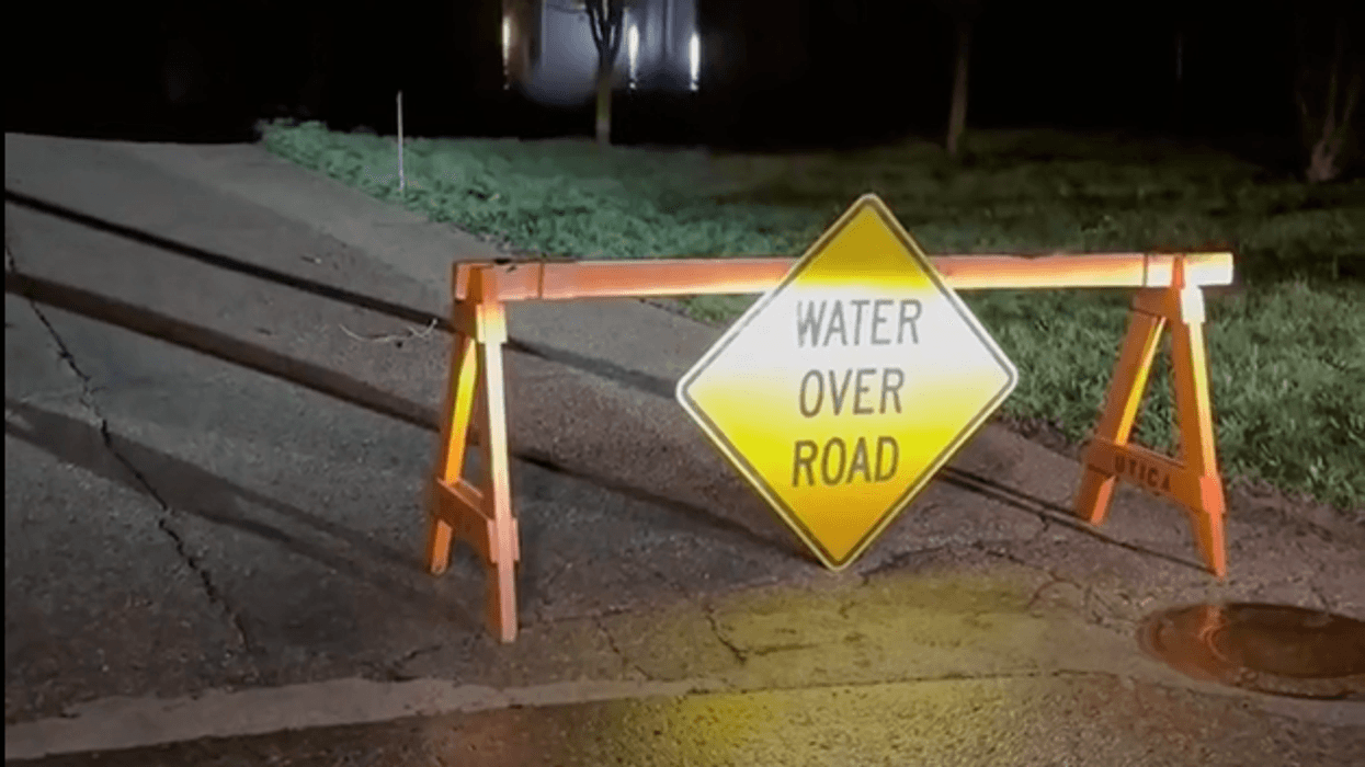 Yellow 'Water Over Road' sign on wooden barrier at night, water reflecting lights nearby.